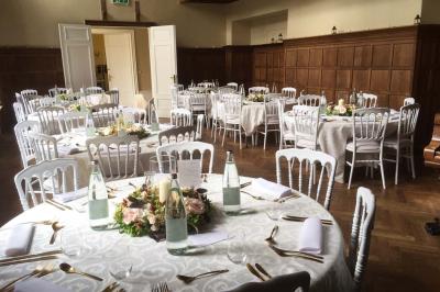 Salle de réception élégante avec tables dressées, chaises blanches, décorations florales et un piano en arrière-plan.