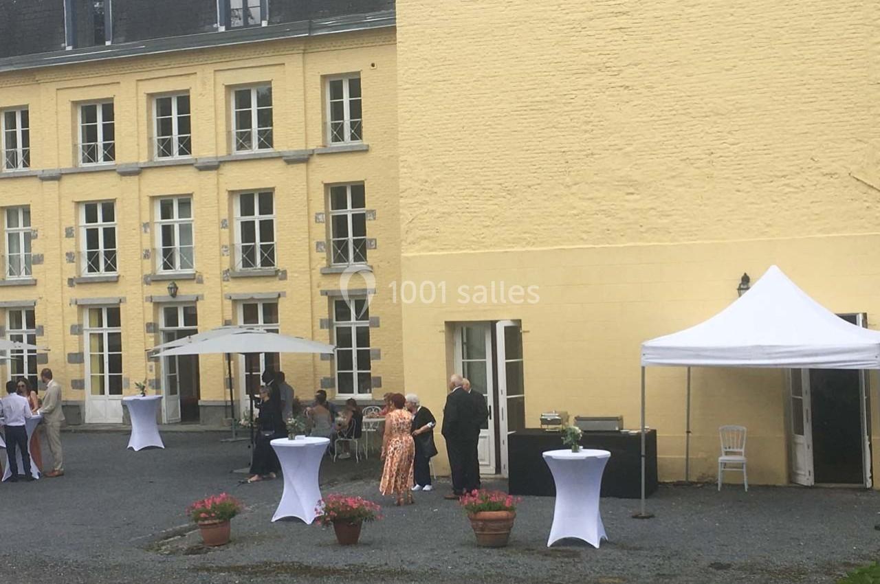 Personnes rassemblées dans une cour pavée devant un bâtiment jaune, avec des tables hautes et des tentes blanches.