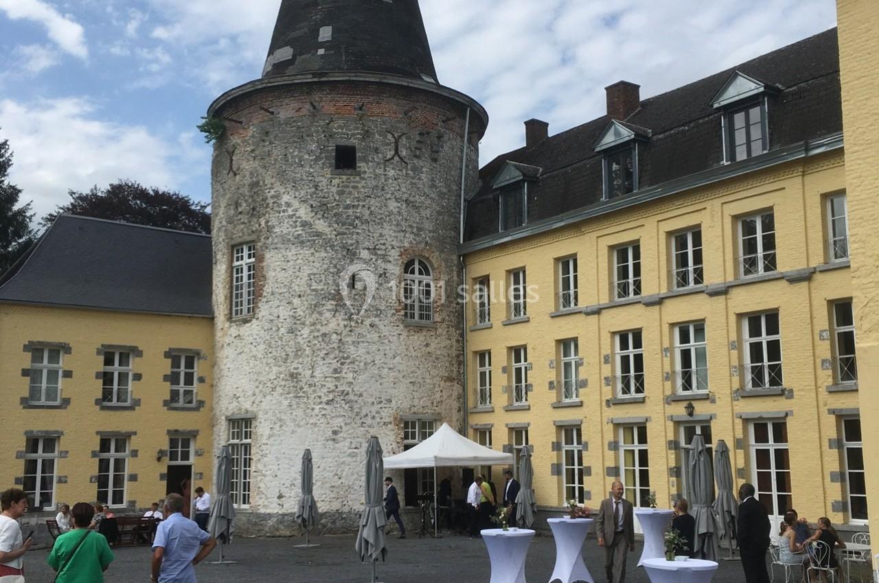 Cour d’un château avec une tour en pierre et des bâtiments jaunes, tables dressées et personnes discutant en extérieur.