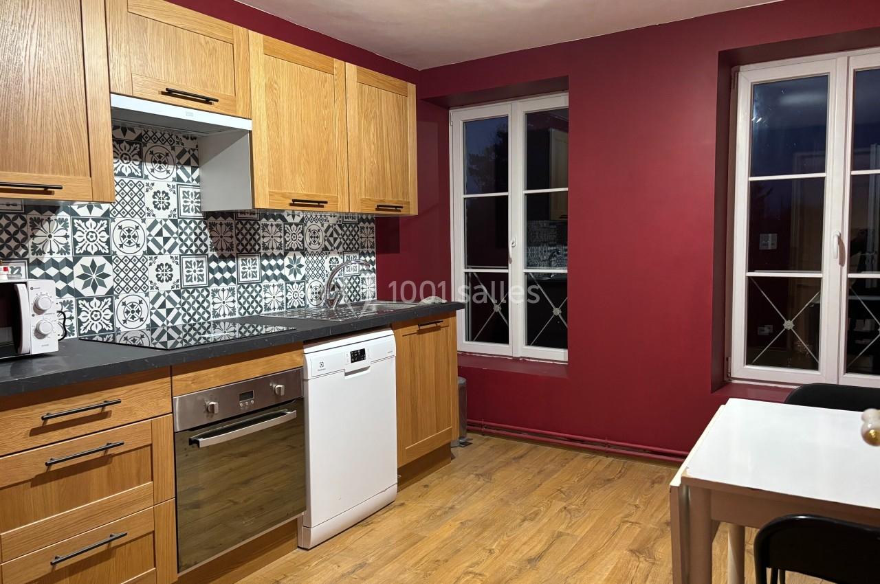 Cuisine avec meubles en bois clair, crédence à motifs, murs rouges, sol en parquet et table blanche près des fenêtres.