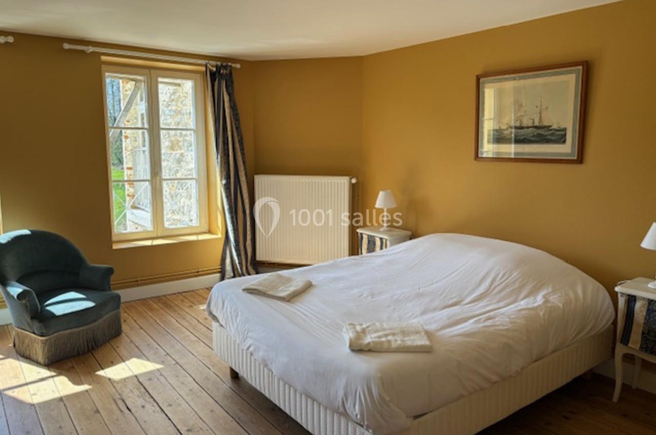Chambre lumineuse avec murs jaunes, lit double, fauteuil, fenêtres donnant sur l'extérieur et parquet en bois.