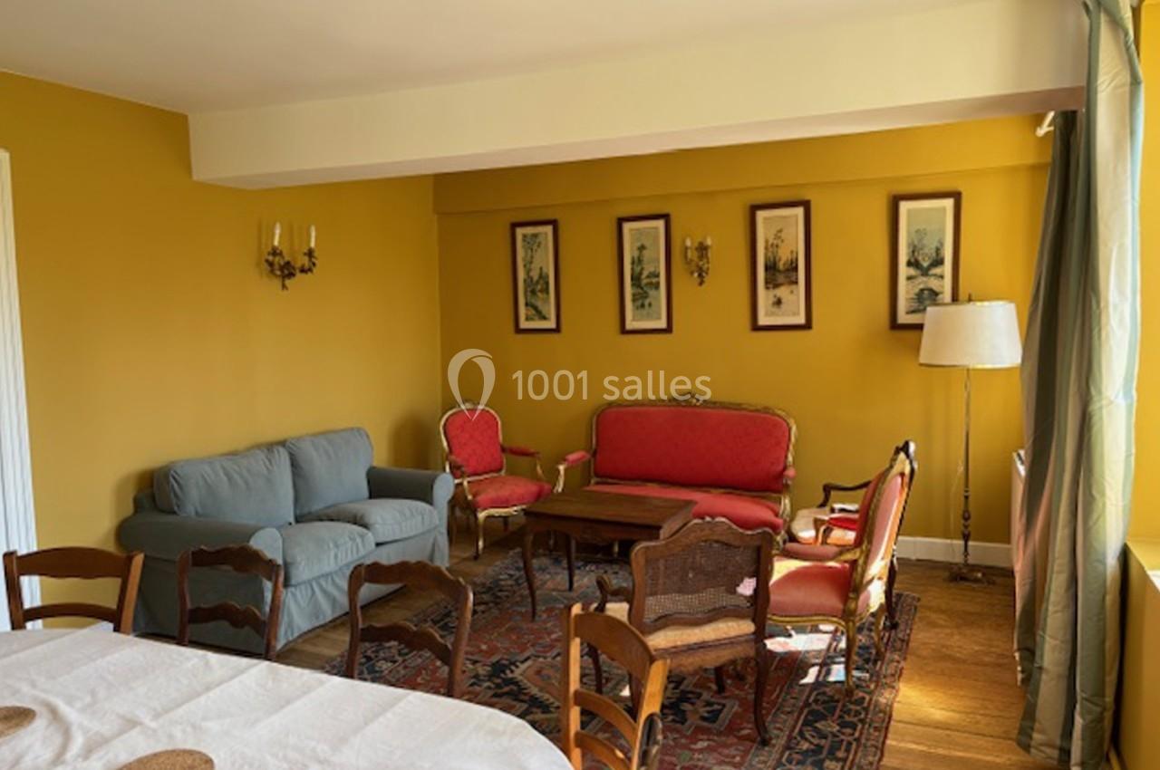 Salon avec murs jaunes, mobilier ancien comprenant canapés, fauteuils rouges, table basse et tapis, éclairé par une fenêtre.