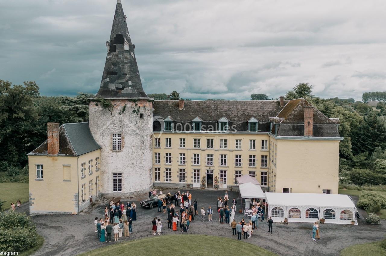 Vue aérienne d'un château avec une tour, une cour animée par des invités et une tente blanche installée à côté.