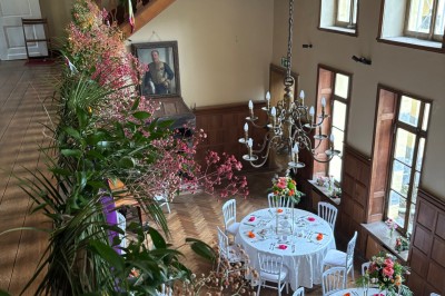 Salle de réception élégante avec tables dressées, chaises blanches, décorations florales et un piano en arrière-plan.