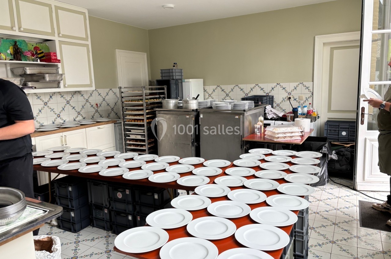 Des assiettes blanches alignées sur des tables dans une cuisine équipée, préparées pour un événement.