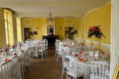 Salle de réception élégante avec tables dressées, chaises blanches, décorations florales et un piano en arrière-plan.