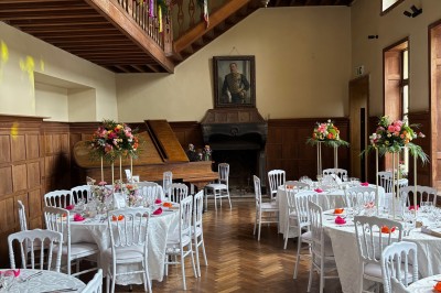 Salle de réception élégante avec tables dressées, chaises blanches, décorations florales et un piano en arrière-plan.