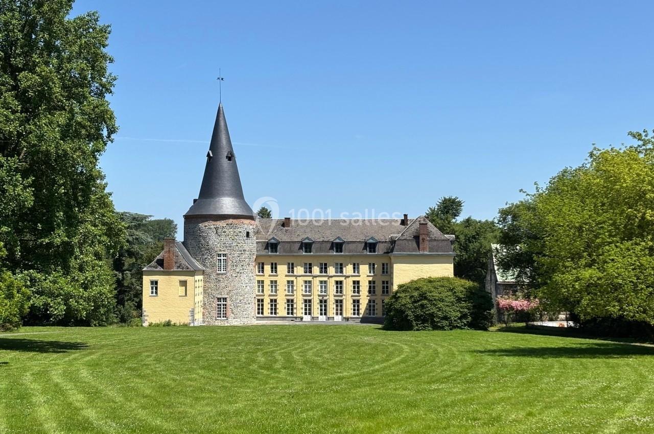 Château jaune avec une tour en pierre et un toit conique, entouré d'une pelouse et d'arbres sous un ciel bleu.