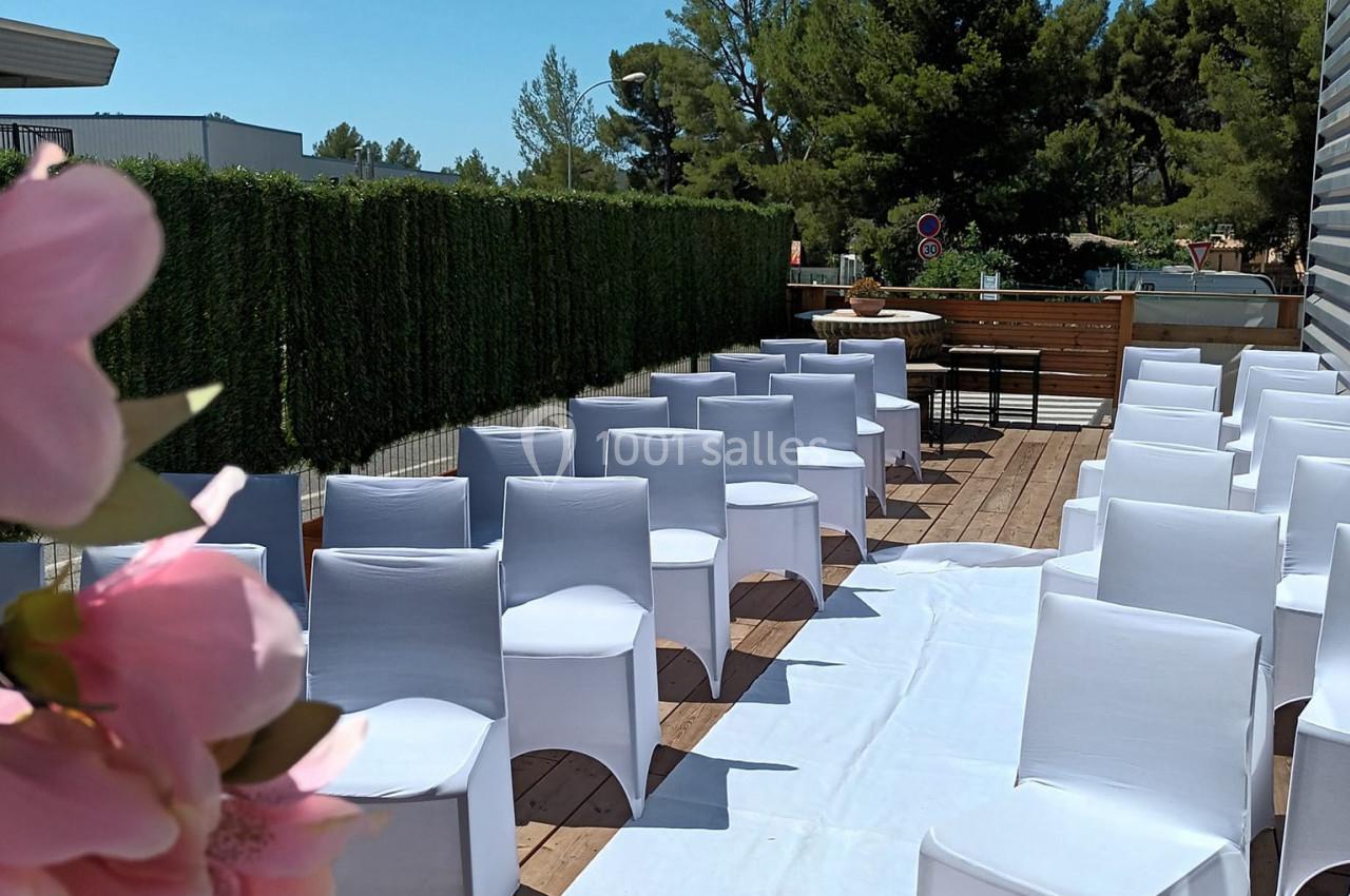 Chaises blanches alignées sur une terrasse en bois, décorée de fleurs roses, sous un ciel bleu clair.