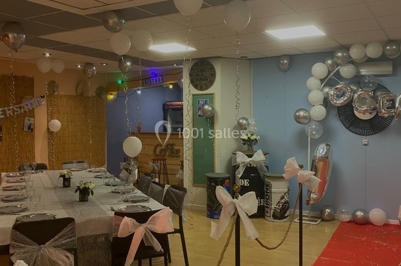 Salle décorée pour une fête avec ballons blancs et argentés, nappes ornées, rubans et tapis rouge.
