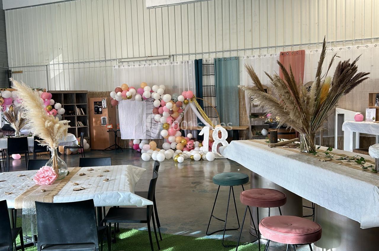Salle décorée pour une fête avec ballons, pampas, tables dressées et le chiffre 18 en grand au centre.