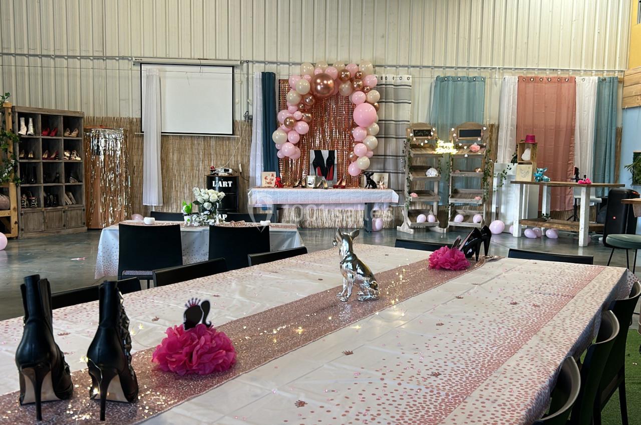 Salle décorée pour une fête avec des tables ornées de nappes pailletées, ballons roses et éléments festifs.