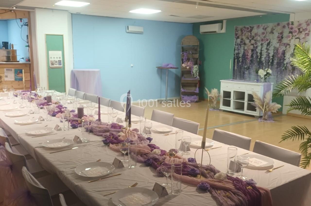 Table décorée pour un événement, ornée de nappes blanches, chemins de table violets et bougies, dans une salle pastel.