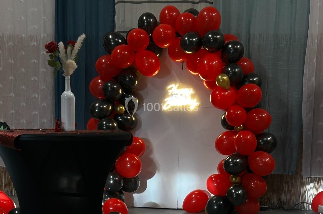 Arche décorative en ballons rouges et noirs avec une inscription lumineuse, table avec vase et fleurs au premier plan.