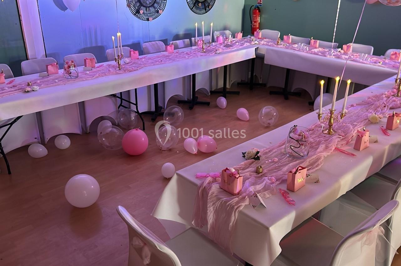 Salle décorée pour une fête, avec tables en U ornées de nappes blanches, bougies, ballons et cadeaux roses.