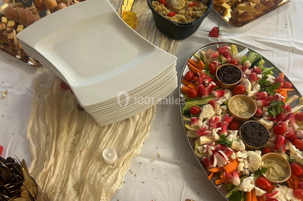 Buffet avec plateaux de légumes, trempettes, pains et amuse-bouches disposés sur une nappe blanche décorée.