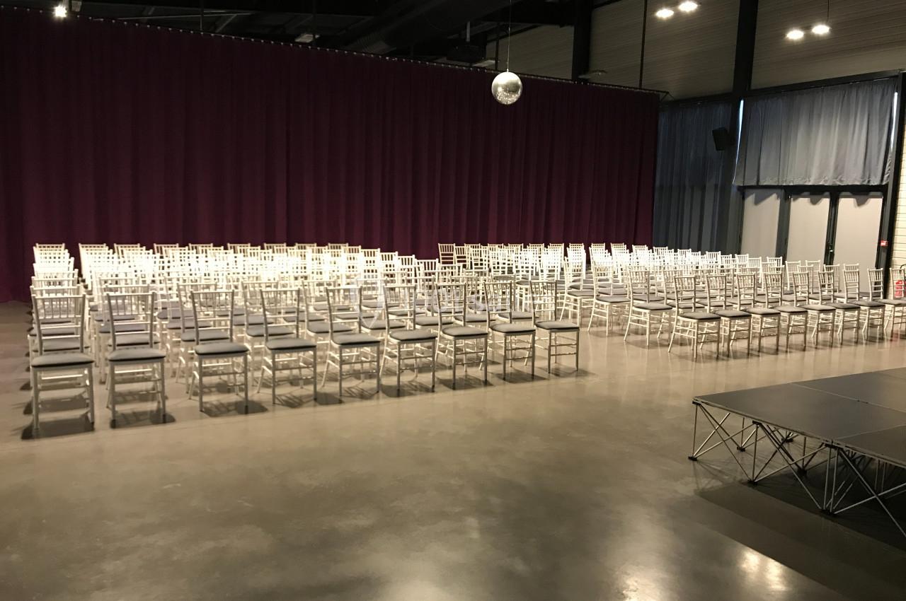 Salle de conférence avec des rangées de chaises blanches alignées devant une scène et un rideau violet.