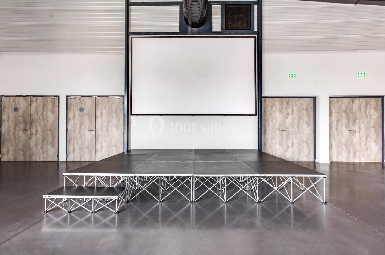 Estrade modulaire en métal avec une petite marche, installée dans une salle avec écran blanc et portes en bois.
