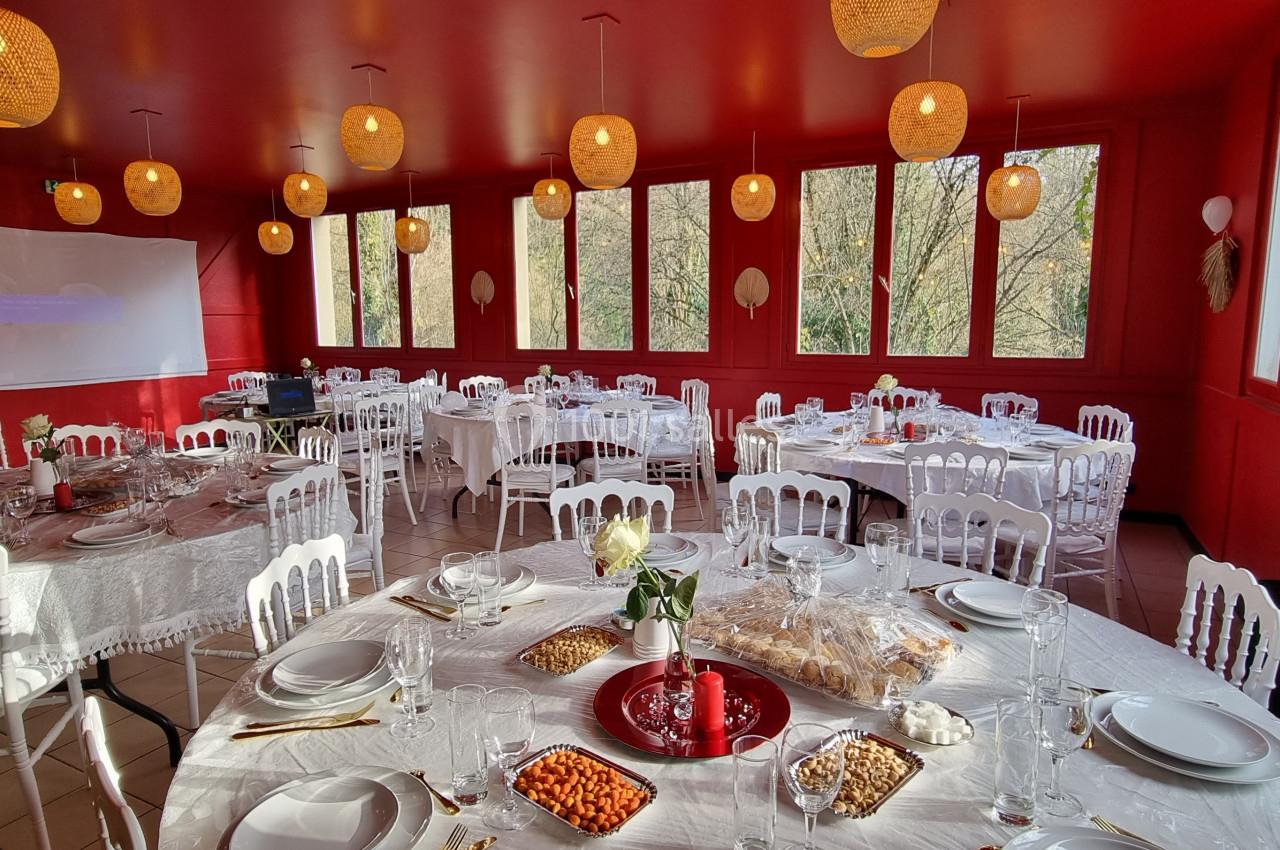 Salle de réception lumineuse avec murs rouges, tables rondes dressées de nappes blanches et décorations suspendues.