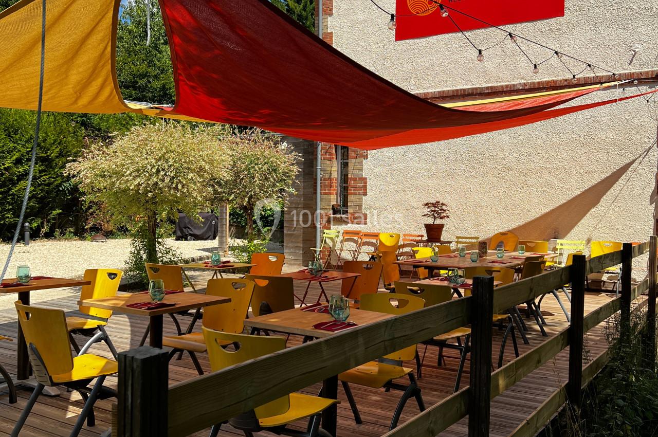 Terrasse en bois avec des tables et chaises jaunes, ombragée par des voiles colorées, devant un bâtiment clair.