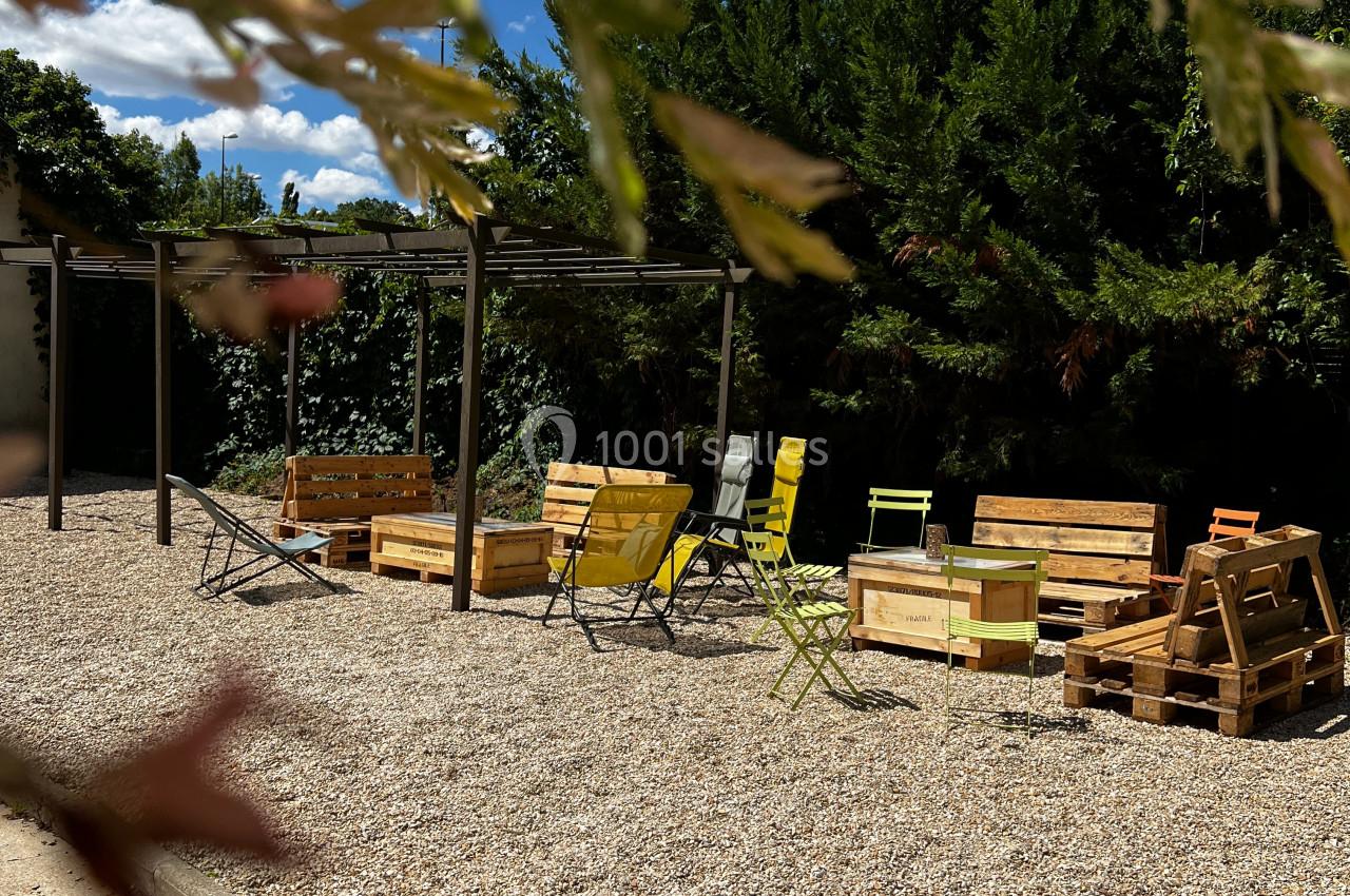 Espace extérieur avec chaises colorées et mobilier en palettes sur un sol gravillonné, entouré de végétation.