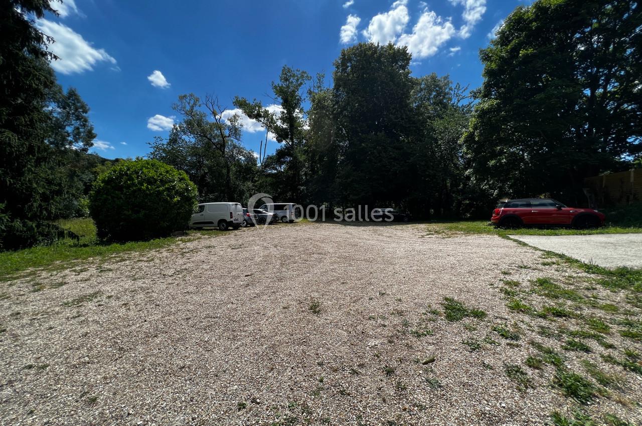Parking extérieur non asphalté avec plusieurs voitures stationnées, entouré d'arbres sous un ciel partiellement nuageux.