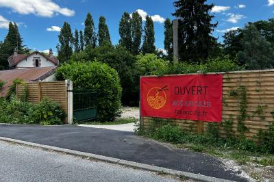 Banderole rouge annonçant l'ouverture d'un café, accrochée à une clôture en bois près d'un portail vert.