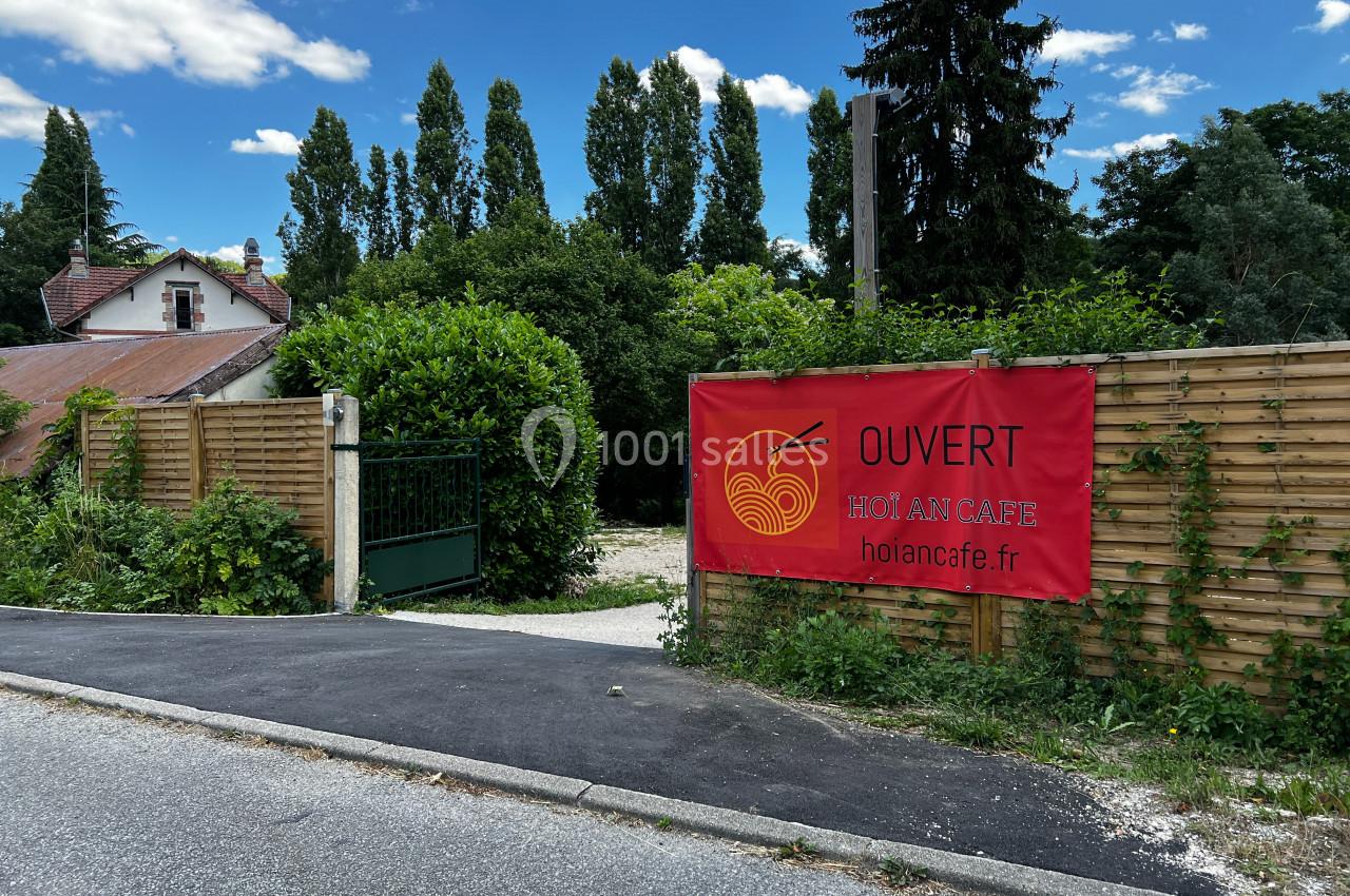 Banderole rouge annonçant l'ouverture d'un café, accrochée à une clôture en bois près d'un portail vert.