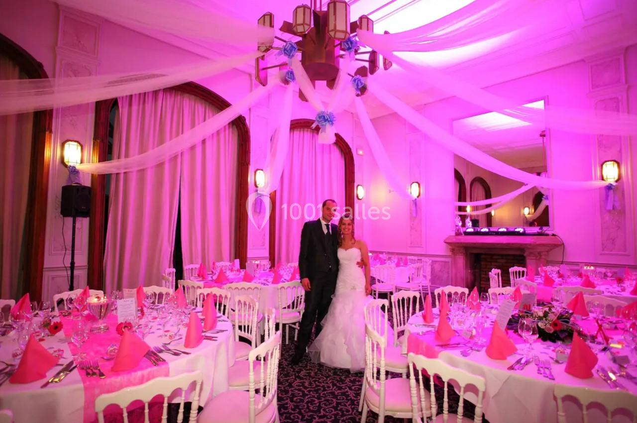 Un couple en tenue de mariage pose dans une salle décorée de nappes roses et de voilages sous un éclairage rose.