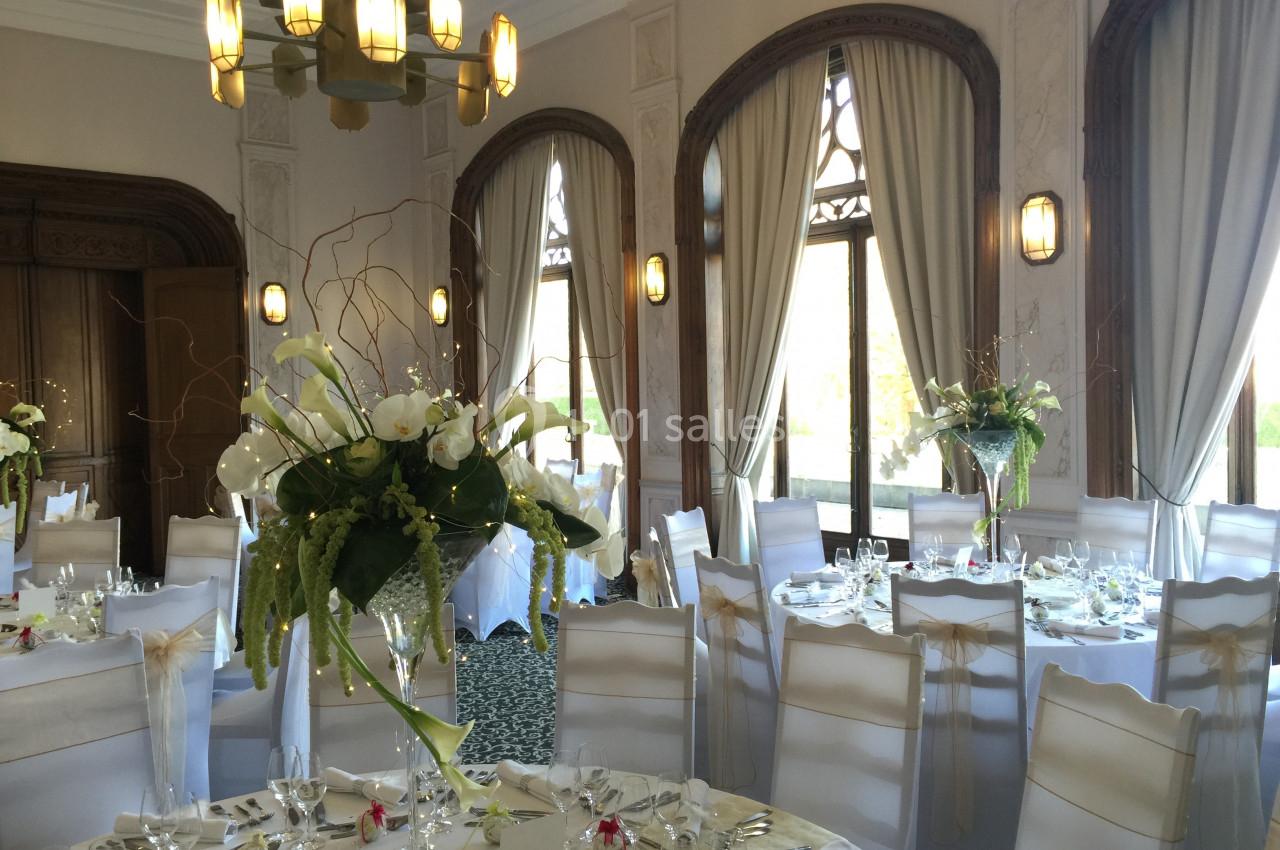 Salle de réception élégante avec tables rondes dressées, chaises blanches et grandes compositions florales.