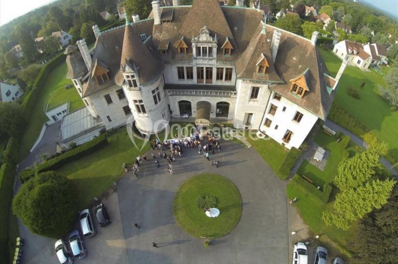 Vue aérienne d'un grand manoir avec une cour centrale où des personnes sont rassemblées, entouré de verdure.