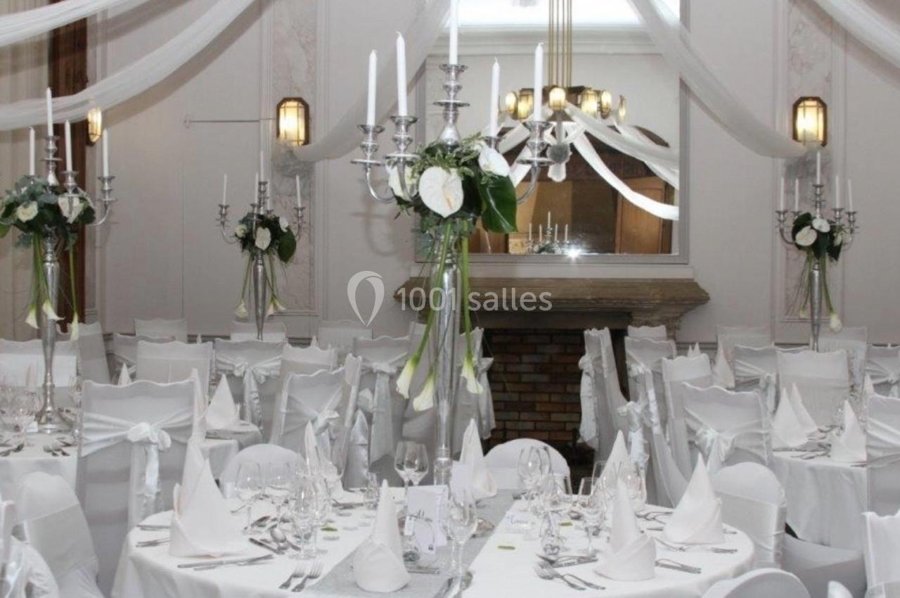 Salle de réception décorée en blanc avec chandeliers, nappes élégantes et compositions florales sur les tables.