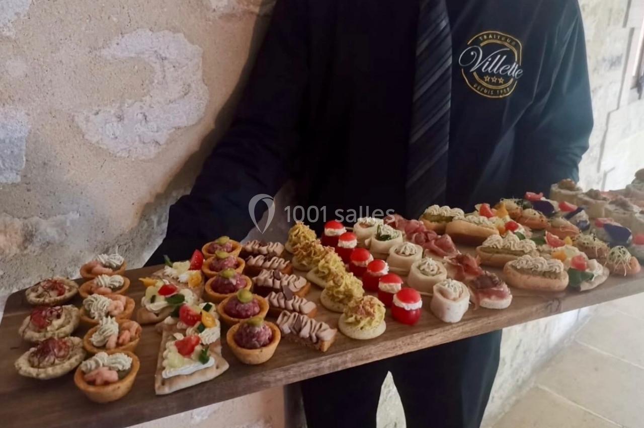 Plateau de canapés variés présentés par un serveur en tenue noire, devant un mur en pierre.