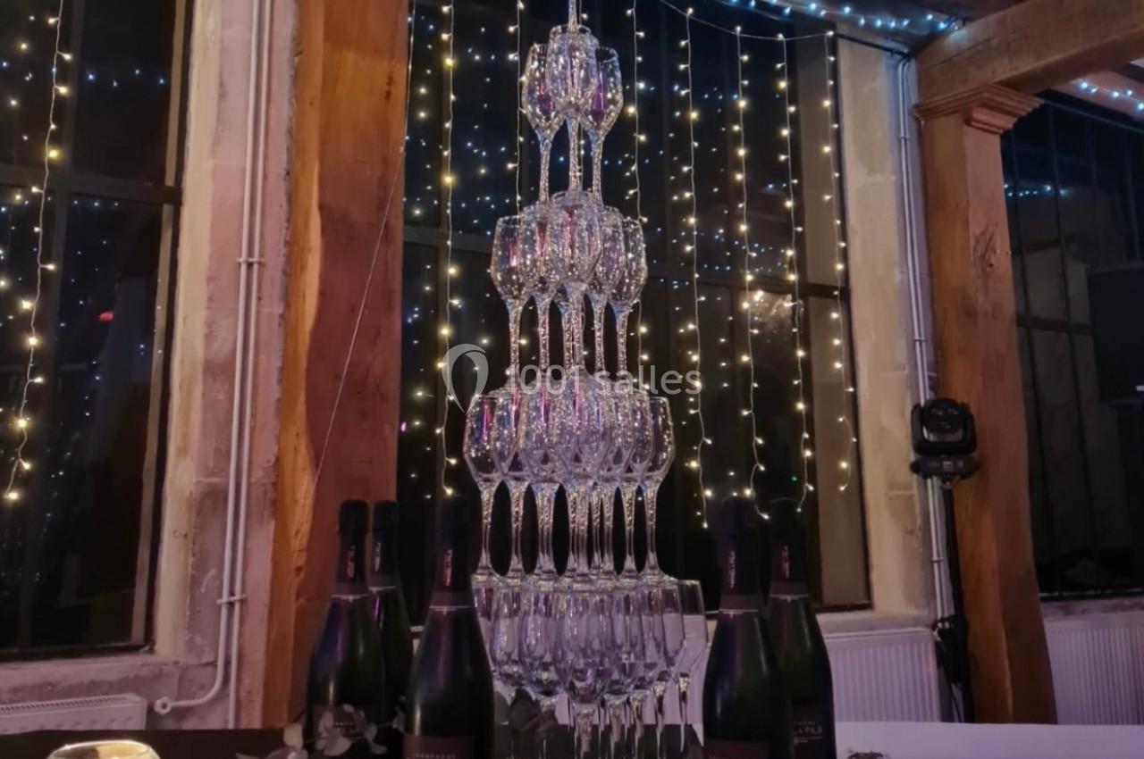 Tour de coupes en verre empilées pour un service de champagne, entourée de bouteilles et de décorations florales.
