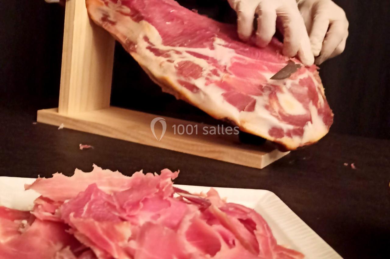 Un serveur découpe finement un jambon sur un support en bois, avec des tranches disposées sur une assiette au premier plan.