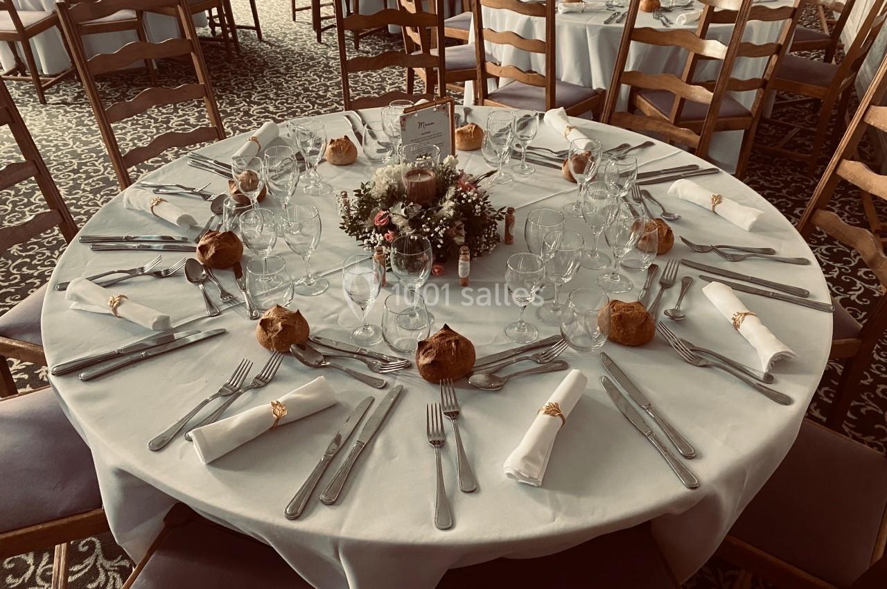 Table ronde dressée pour un repas, avec nappes blanches, couverts, verres, serviettes et décoration florale centrale.