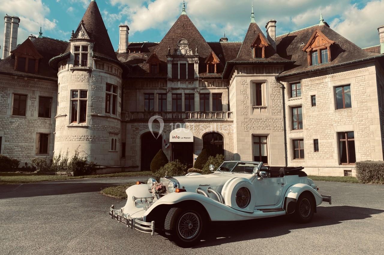 Voiture ancienne blanche garée devant un château en pierre avec des toits en ardoise sous un ciel partiellement nuageux.