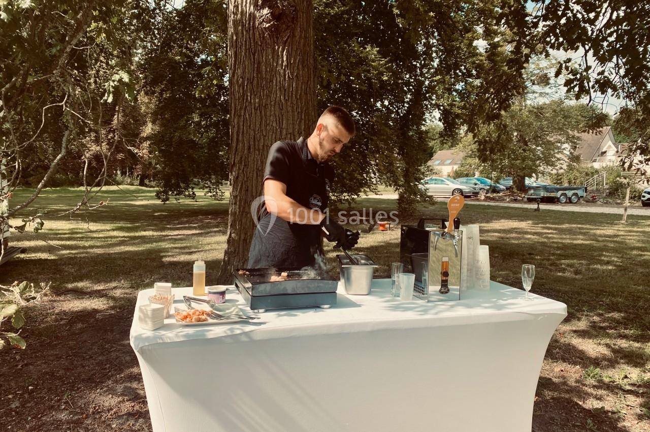 Un cuisinier prépare des plats sur une table blanche en extérieur, entouré d'arbres et d'herbe.