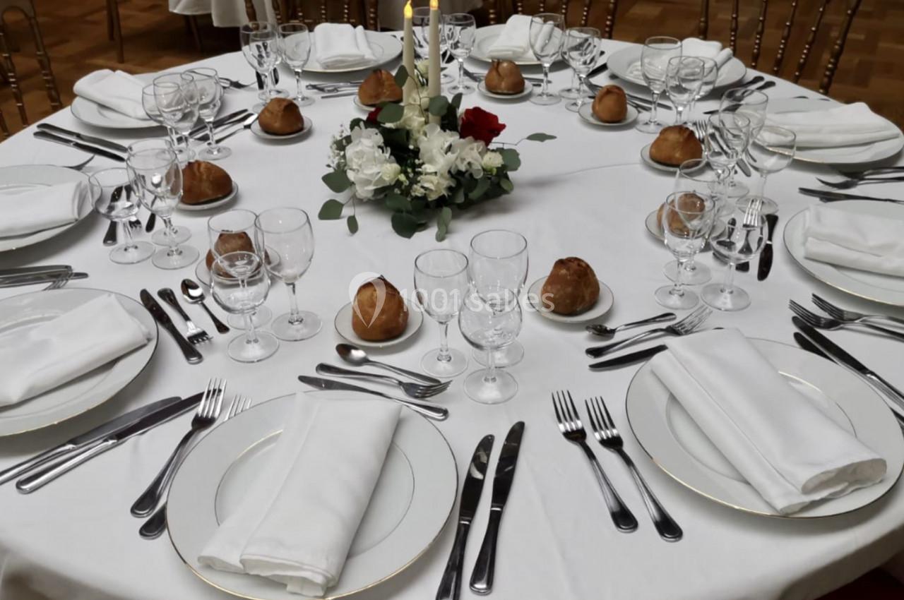 Table ronde élégamment dressée avec nappes blanches, couverts, verres, petits pains et centre de table floral.