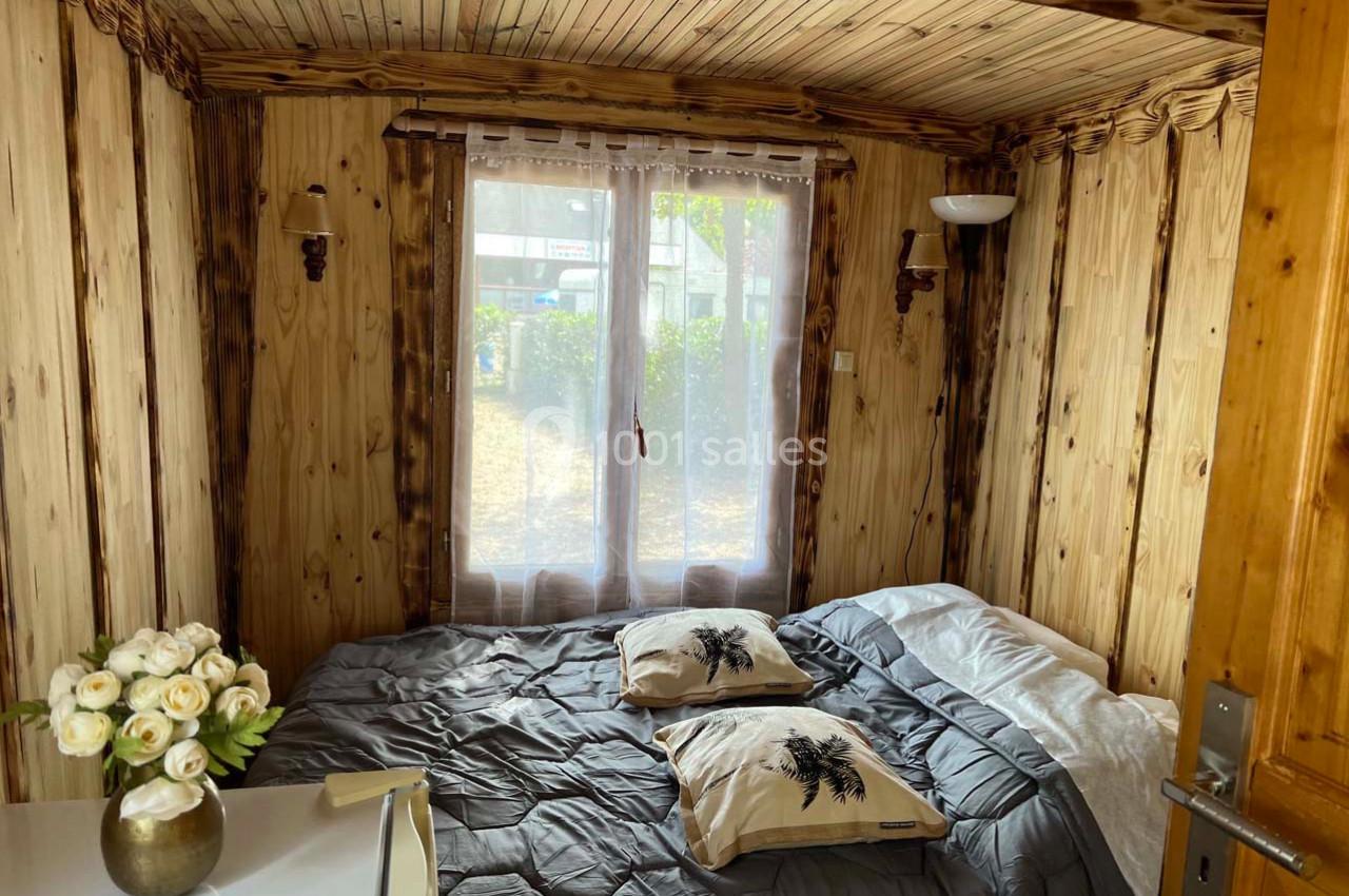 Petite chambre en bois avec un lit double, des coussins décoratifs, une fenêtre avec rideaux et un bouquet de fleurs.