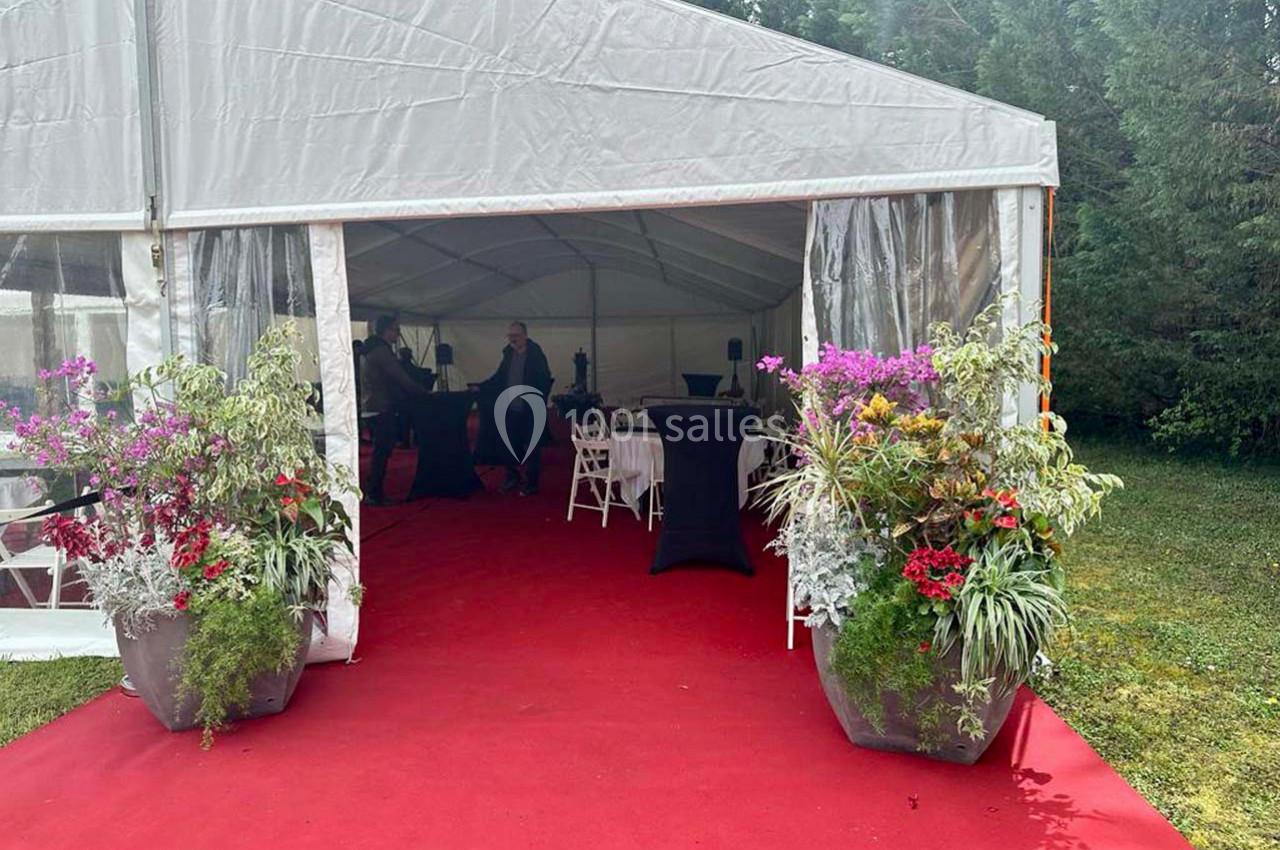 Entrée d'une tente événementielle avec tapis rouge, grandes plantes fleuries et mobilier de réception à l'intérieur.