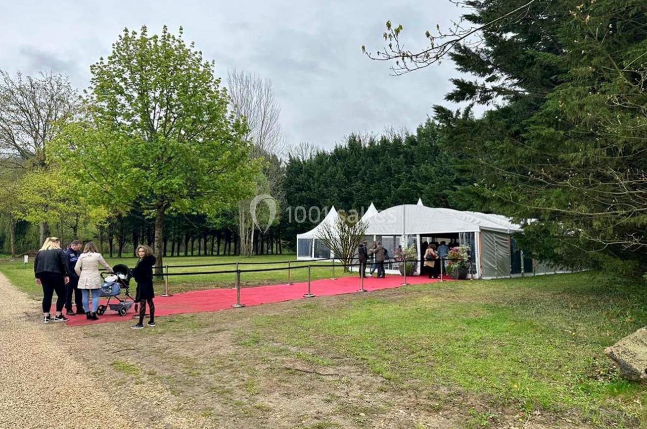 Tente blanche avec tapis rouge menant à l'entrée, entourée d'arbres, avec des visiteurs arrivant.