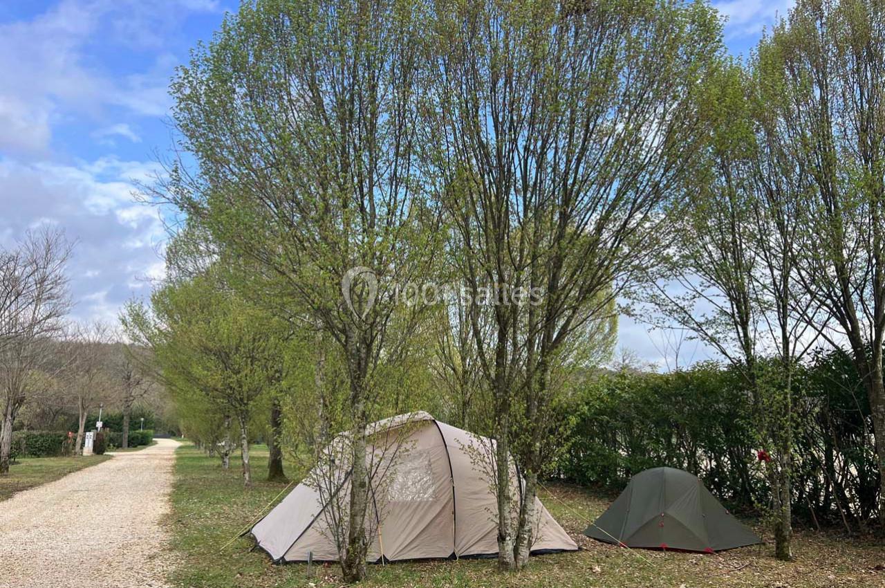 Deux tentes installées sous des arbres dans un camping, avec un chemin de gravier à gauche et un ciel partiellement nuageux.
