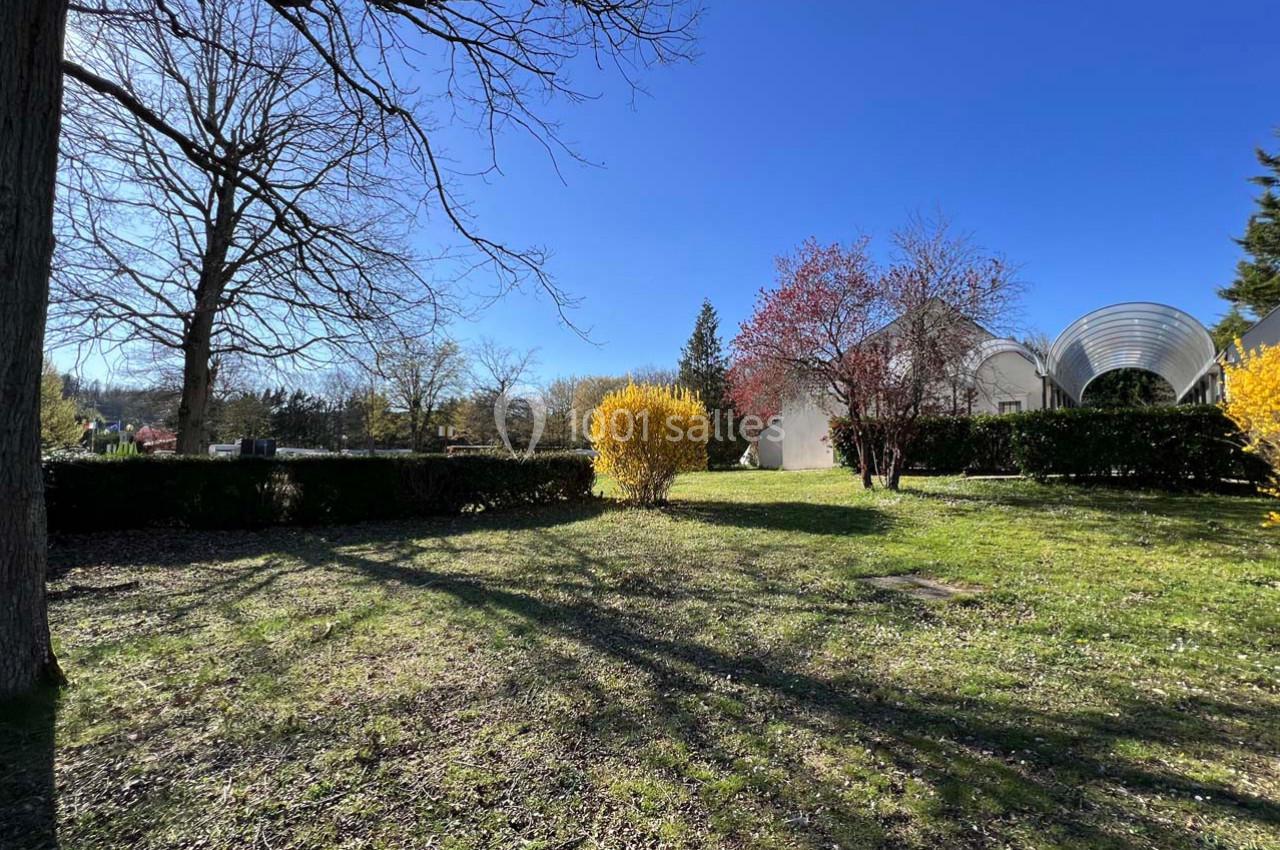 Jardin ensoleillé avec pelouse, arbres dénudés, arbustes fleuris et bâtiment blanc en arrière-plan.