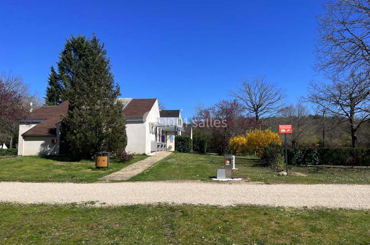 Maison blanche avec toit brun entourée de végétation, chemin en gravier et panneau rouge visible au premier plan.