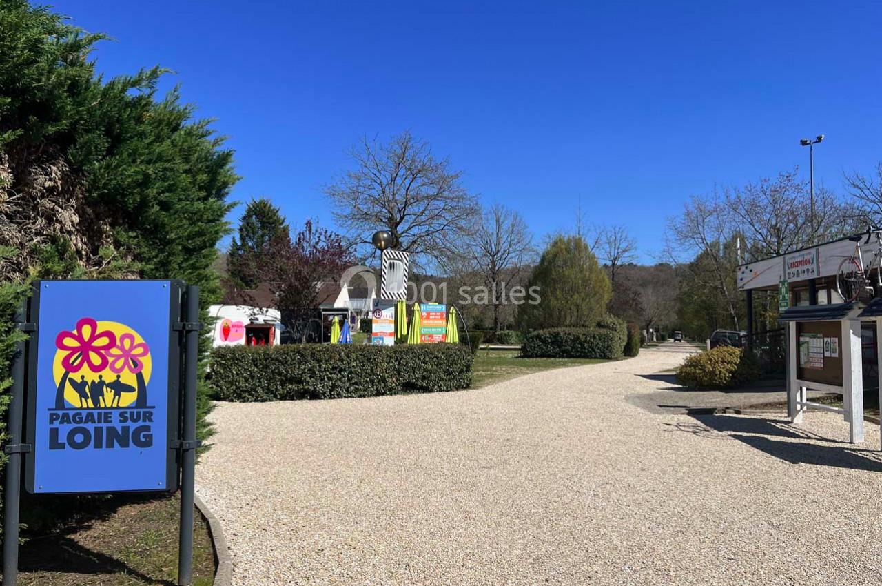 Chemin gravillonné bordé de verdure avec un panneau indiquant ’Pagaie sur Loing’ et des installations colorées au fond.