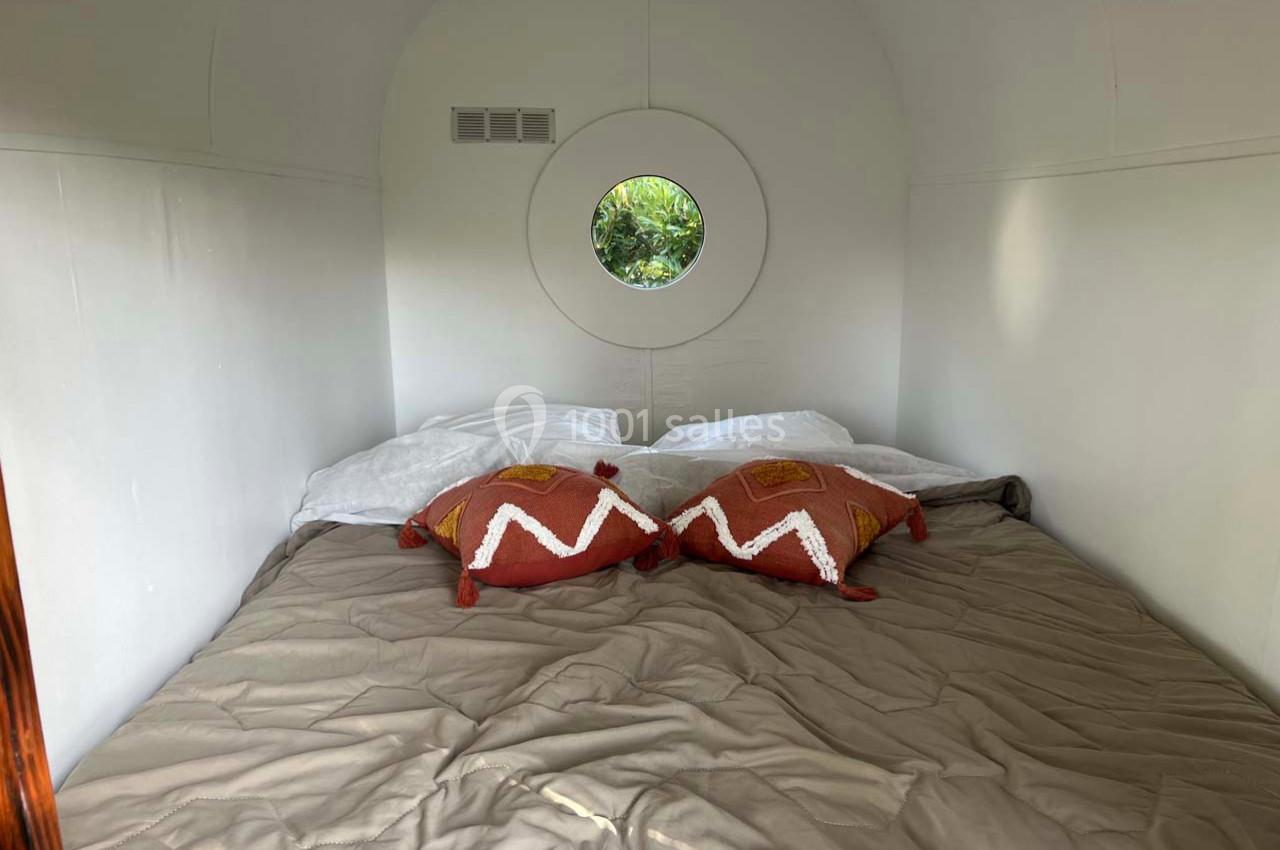 Lit double avec couverture beige, deux coussins décoratifs rouges et fenêtre ronde donnant sur un jardin.