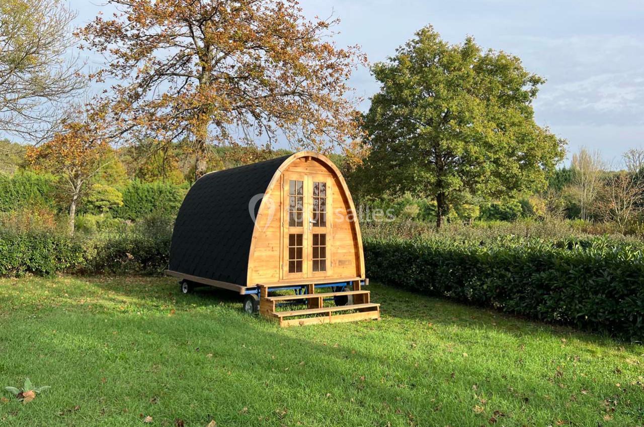Cabane en bois de forme arrondie avec toit sombre, située sur une pelouse entourée d'arbres et de haies.