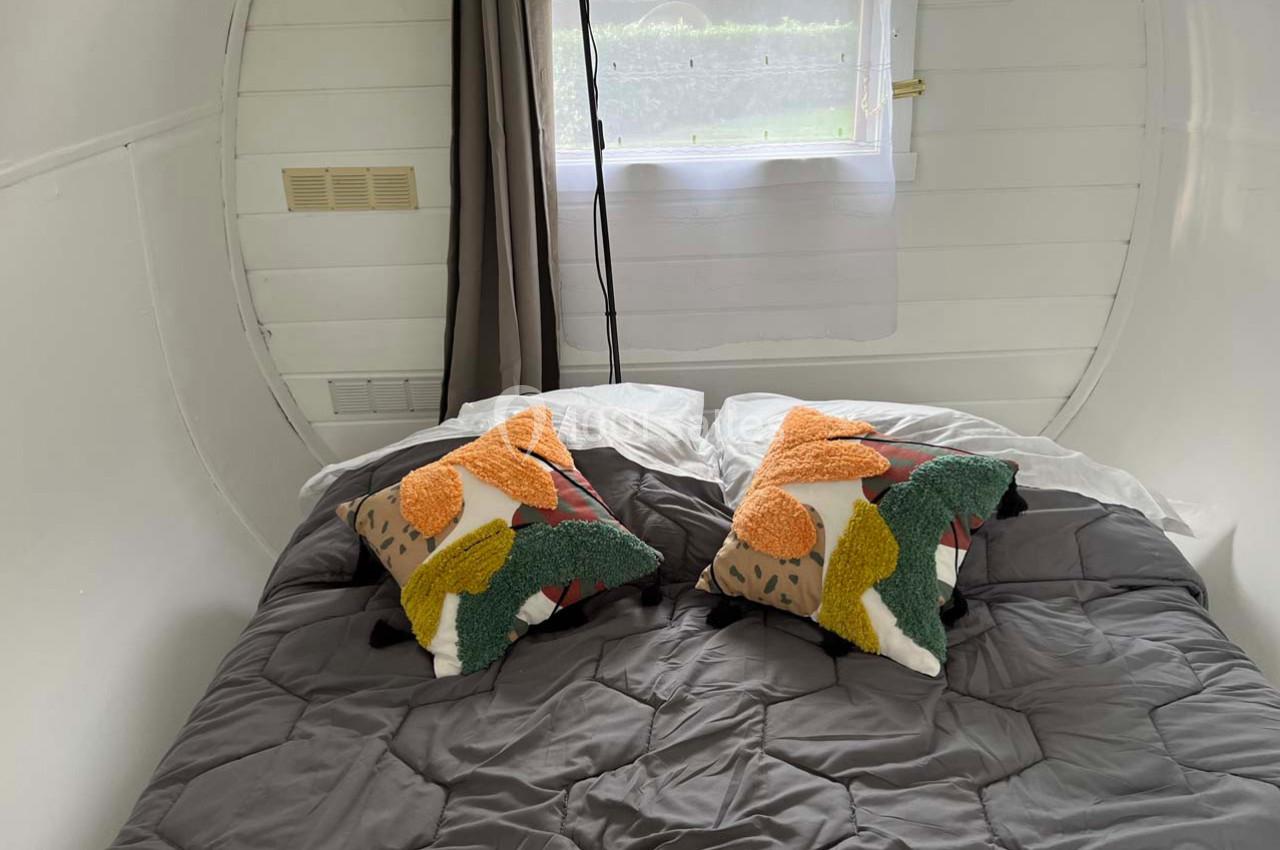 Lit double avec couette grise, deux coussins colorés et une fenêtre avec rideaux dans une pièce blanche.