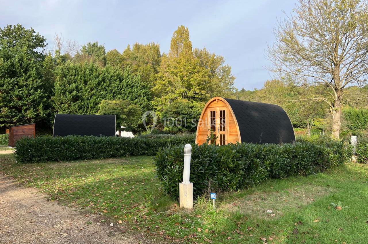 Deux cabanes en bois en forme d'arc entourées de haies, situées dans un espace vert arboré.